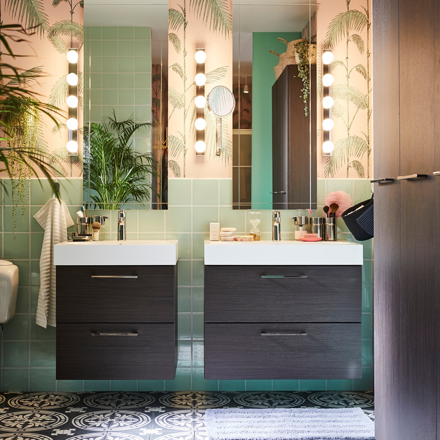 Bathroom with two mirrors, two sink and storage units with diverse toiletry articles plus wall lighting and a large plant.
