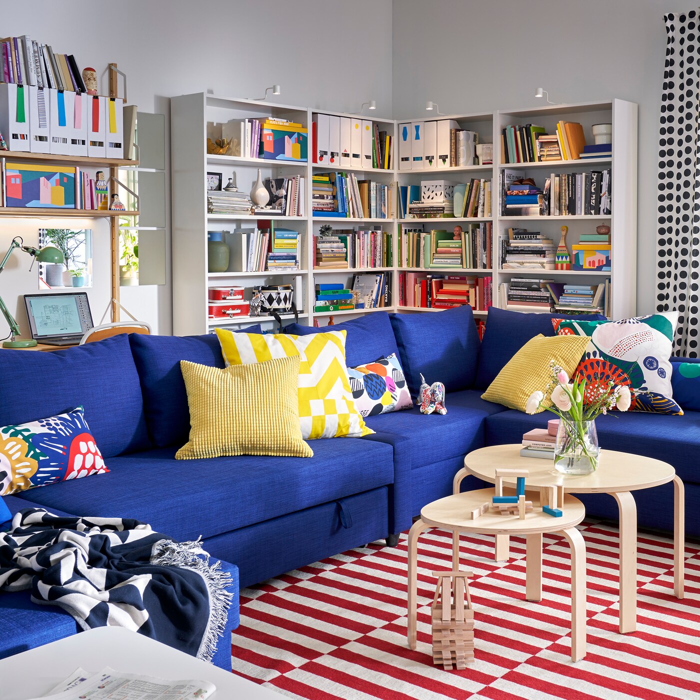 Living room with FRIHETEN blue corner sofa bed with lots of pillows, some bookcases and a window with plants in the sill.